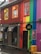 A vibrant building features a rainbow-colored facade with stripes of red, orange, yellow, green, blue, and purple. The upper windows have bright orange and yellow frames. There is a sign for 'Kiki queer bar' above the entrance, and several black awnings cover the windows below. The door is painted in black and a sidewalk with a rainy surface is in front of the building. A wooden box is placed near the entrance.