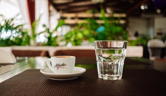 A cup of espresso labeled Alfredo sits next to a glass of water on a brown placemat. The setting appears to be a cozy caf&eacute; with lush green plants in the blurry background, creating a relaxed and inviting atmosphere.