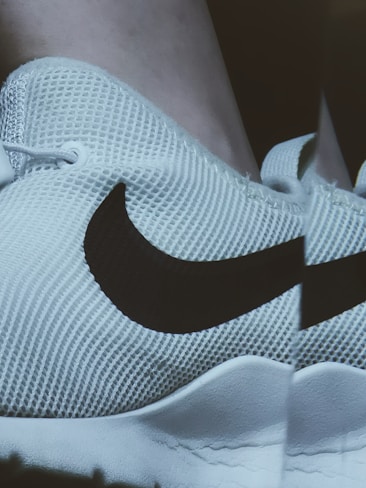 A close-up view of a white sneaker with a prominent black logo on the side. The shoe has a mesh texture and a cushioned sole.