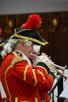 A person dressed in an elaborate historical uniform plays a trumpet. The uniform is red with gold trim and features a feathered hat, alongside white curly hair characteristic of historical wigs. The setting appears to be an indoor performance, possibly involving a group.