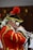 A person dressed in an elaborate historical uniform plays a trumpet. The uniform is red with gold trim and features a feathered hat, alongside white curly hair characteristic of historical wigs. The setting appears to be an indoor performance, possibly involving a group.