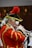 A person dressed in an elaborate historical uniform plays a trumpet. The uniform is red with gold trim and features a feathered hat, alongside white curly hair characteristic of historical wigs. The setting appears to be an indoor performance, possibly involving a group.
