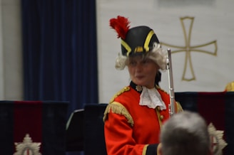 A person is dressed in historical military attire, featuring a red coat with gold accents and a black hat adorned with a red plume. They are holding a silver ceremonial staff with an ornate cross in the background.