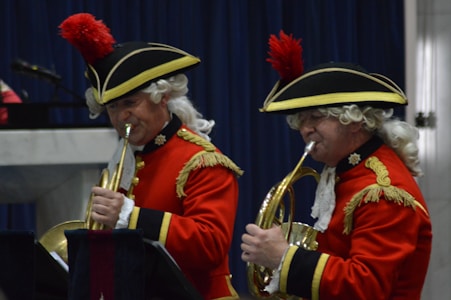 Two musicians dressed in historical military uniforms are playing musical instruments. They are wearing red coats with gold embroidery and black hats adorned with red plumes. The musicians have white wigs and are focused on their performance.