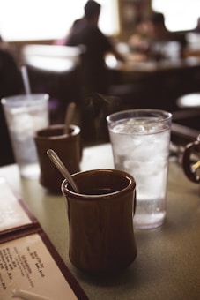 A cozy coffee shop setting with a focus on a brown ceramic mug filled with hot coffee, accompanied by a spoon. Nearby, there is a clear plastic glass filled with ice water. The background is softly blurred, featuring silhouettes of patrons sitting at tables, creating a warm and inviting ambiance.