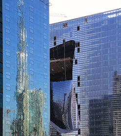 A modern urban landscape featuring high-rise buildings with glass facades. The reflections of the surrounding architecture are visible on the windows, creating a distorted and dynamic visual effect. A construction crane can be seen in the background, suggesting ongoing development in the area.