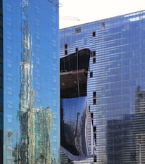 A modern urban landscape featuring high-rise buildings with glass facades. The reflections of the surrounding architecture are visible on the windows, creating a distorted and dynamic visual effect. A construction crane can be seen in the background, suggesting ongoing development in the area.