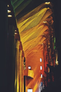 A vibrant interior with dynamic architectural structures featuring sharp, angular lines and high ceilings. Warm lighting highlights the intricate patterns on the surfaces, blending shades of gold, yellow, and orange. Small rectangular windows are scattered across the walls.