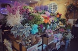 A vibrant floral shop display featuring a wide variety of colorful flowers arranged in vases and wrappings. The arrangement includes shades of pink, purple, orange, and white flowers, surrounded by lush green foliage. The setting is warm with exposed brick walls and decorative elements like wreaths and a neon sign.