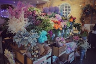 A vibrant floral shop display featuring a wide variety of colorful flowers arranged in vases and wrappings. The arrangement includes shades of pink, purple, orange, and white flowers, surrounded by lush green foliage. The setting is warm with exposed brick walls and decorative elements like wreaths and a neon sign.