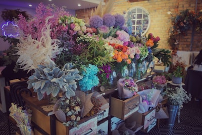 A vibrant floral shop display featuring a wide variety of colorful flowers arranged in vases and wrappings. The arrangement includes shades of pink, purple, orange, and white flowers, surrounded by lush green foliage. The setting is warm with exposed brick walls and decorative elements like wreaths and a neon sign.