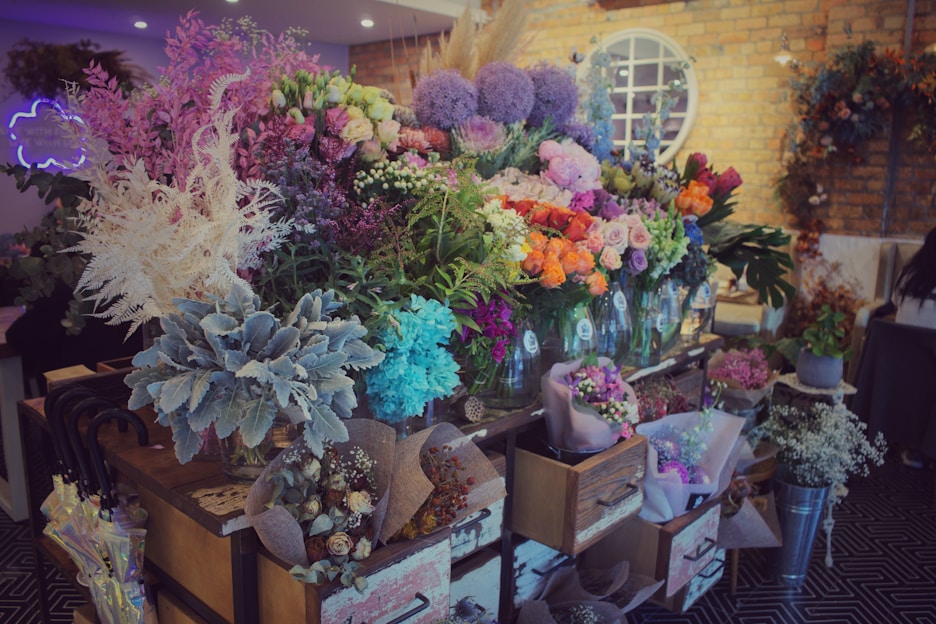 A vibrant floral shop display featuring a wide variety of colorful flowers arranged in vases and wrappings. The arrangement includes shades of pink, purple, orange, and white flowers, surrounded by lush green foliage. The setting is warm with exposed brick walls and decorative elements like wreaths and a neon sign.