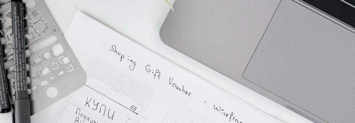 A well-organized workspace featuring a laptop, a potted plant, design sketches, and writing tools. The sketches appear to be wireframes for a 'Shopping Gift Voucher' application or website, drawn on white paper with annotations.