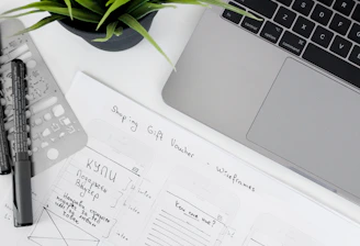 A well-organized workspace featuring a laptop, a potted plant, design sketches, and writing tools. The sketches appear to be wireframes for a 'Shopping Gift Voucher' application or website, drawn on white paper with annotations.
