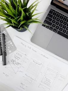 A well-organized workspace featuring a laptop, a potted plant, design sketches, and writing tools. The sketches appear to be wireframes for a 'Shopping Gift Voucher' application or website, drawn on white paper with annotations.