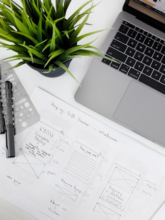 A well-organized workspace featuring a laptop, a potted plant, design sketches, and writing tools. The sketches appear to be wireframes for a 'Shopping Gift Voucher' application or website, drawn on white paper with annotations.