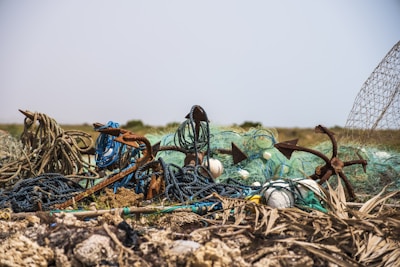 A pile of discarded fishing nets and anchors lies on a rocky and sandy terrain. The nets are tangled and weathered, with some ropes appearing frayed and rusted. The background is an arid, open landscape under a clear sky.