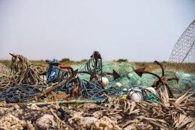 A pile of discarded fishing nets and anchors lies on a rocky and sandy terrain. The nets are tangled and weathered, with some ropes appearing frayed and rusted. The background is an arid, open landscape under a clear sky.