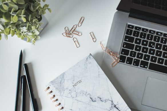 A neatly arranged workspace features an open laptop with a visible keyboard. A marble-patterned notebook labeled 'NOTES' lies nearby. Several copper paper clips are scattered across the surface, adding a metallic touch. Two sleek black pens rest close to the notebook, suggesting readiness for writing. A lush green plant in a white pot adds a natural element to the otherwise modern setup.