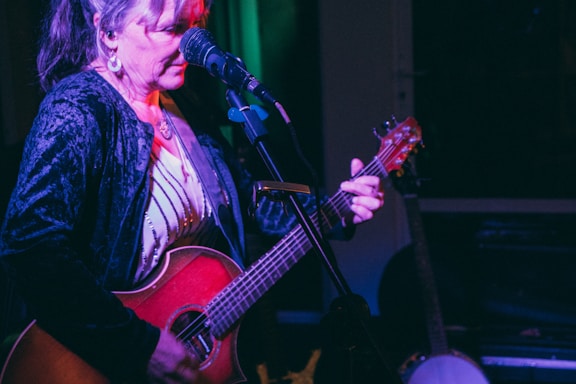 A woman is passionately performing with an acoustic guitar on stage. She has long hair tied back, and is focused on her music. She wears a black velvet jacket and stands in front of a microphone, with dim lighting providing a warm, intimate atmosphere. A banjo is visible in the background, suggesting a musical setting.