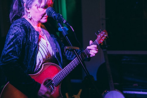A woman is passionately performing with an acoustic guitar on stage. She has long hair tied back, and is focused on her music. She wears a black velvet jacket and stands in front of a microphone, with dim lighting providing a warm, intimate atmosphere. A banjo is visible in the background, suggesting a musical setting.