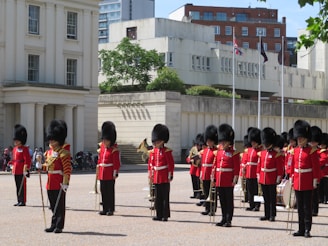 A group of uniformed guardsmen in red tunics and bearskin hats stand in formation on a parade ground. They hold musical instruments and are positioned in front of a large building with white columns. Flags can be seen on poles in the background.