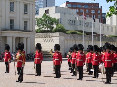 A group of uniformed guardsmen in red tunics and bearskin hats stand in formation on a parade ground. They hold musical instruments and are positioned in front of a large building with white columns. Flags can be seen on poles in the background.