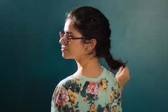 A person with braided hair is seen in profile, wearing glasses and a floral-patterned top with colorful flowers. The background is a solid teal color, creating a contrast with the subject.
