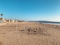 A sandy beach with multiple volleyball courts set up. Several people are playing volleyball, while others relax or walk on the beach. Residential homes and palm trees line the background, and the ocean is visible to the right under a clear blue sky.