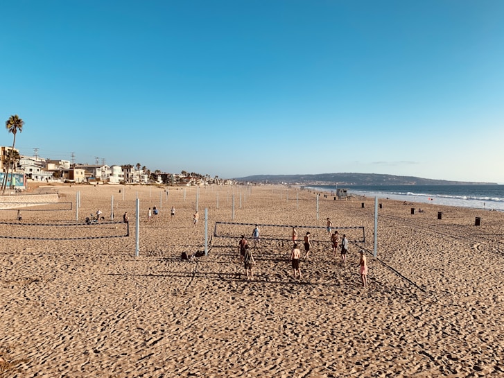 A sandy beach with multiple volleyball courts set up. Several people are playing volleyball, while others relax or walk on the beach. Residential homes and palm trees line the background, and the ocean is visible to the right under a clear blue sky.