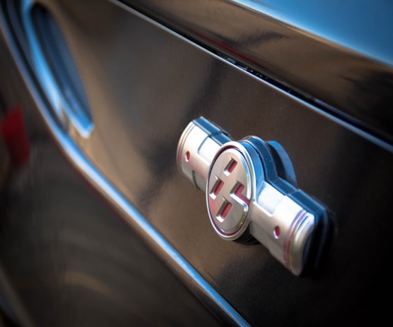 A close-up of a shiny, metallic car emblem with red accents on a dark, reflective surface. The lighting highlights the contours and smooth texture of the badge set against the sleek body of the vehicle.