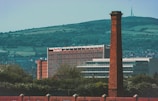 A large brick chimney stands prominently in the foreground with a modern building in the background, which has a sign indicating 'Royal Mail'. The scene also includes rolling green hills and smaller buildings scattered in the distance, against a clear blue sky.