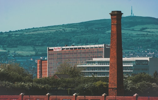 A large brick chimney stands prominently in the foreground with a modern building in the background, which has a sign indicating 'Royal Mail'. The scene also includes rolling green hills and smaller buildings scattered in the distance, against a clear blue sky.