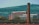 A large brick chimney stands prominently in the foreground with a modern building in the background, which has a sign indicating 'Royal Mail'. The scene also includes rolling green hills and smaller buildings scattered in the distance, against a clear blue sky.