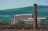 A large brick chimney stands prominently in the foreground with a modern building in the background, which has a sign indicating 'Royal Mail'. The scene also includes rolling green hills and smaller buildings scattered in the distance, against a clear blue sky.