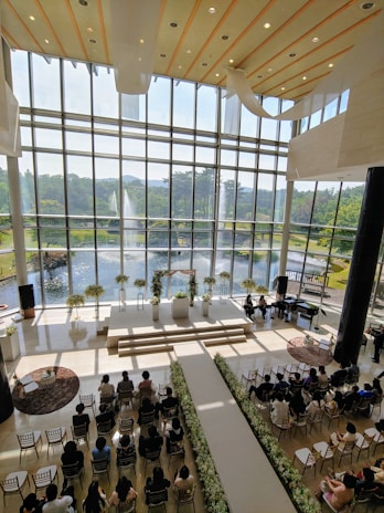 A large indoor venue with a high ceiling and expansive windows overlooking a picturesque garden and fountain outside. The interior features a long aisle adorned with flowers leading up to a stage with floral arrangements. Rows of guests are seated in chairs on either side of the aisle, suggestive of a formal ceremony or event.