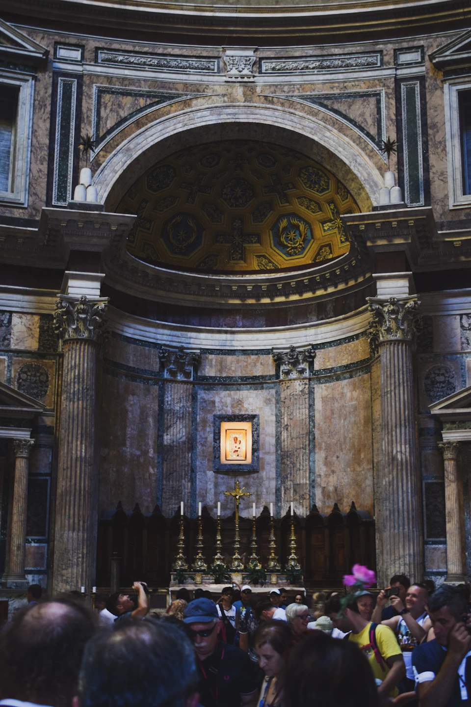 The image depicts a large, ornate room with a high ceiling and intricate decorations. The walls are adorned with gold and white patterns that add to the grandeur of the space. At the center of the room is an altar, which features a cross at its base and a painting above it. A group of people can be seen gathered around the altar, some standing in front of it while others are scattered throughout the room. The overall atmosphere suggests a place of worship or a significant event taking place within this historic building.