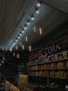 Inside a dimly lit store with a rustic, wooden ceiling, there are several large, globe-shaped hanging light bulbs. A display of various popcorn flavors is lined against the wall, with decorative letters spelling 'CARAMEL CORN' above it. Tins and clear containers filled with popcorn are prominently featured.