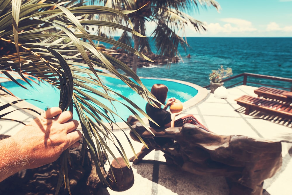 Frau sitzt und hält Kokosnuss in brauner Holzbank in der Nähe des Swimmingpools mit Blick auf das ruhige Meer