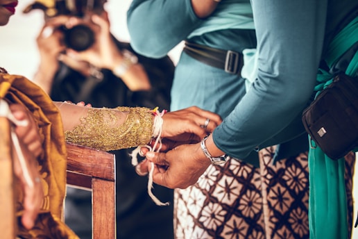 A close-up of a person tying a string around another person's wrist. The person receiving the string has an ornate, gold-colored sleeve. Traditional patterned clothing and colorful fabrics are visible, and a photographer is present in the background capturing the moment.