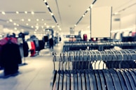 A clothing store interior featuring rows of metal clothing racks filled with hangers. The background is softly blurred, showcasing more clothing displays and bright ceiling lights, creating a spacious and modern retail environment.