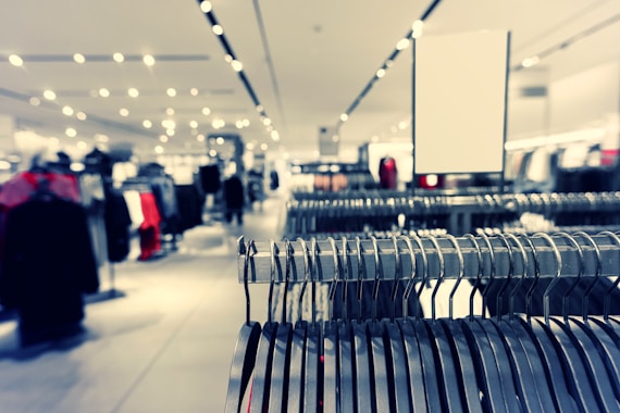 A clothing store interior featuring rows of metal clothing racks filled with hangers. The background is softly blurred, showcasing more clothing displays and bright ceiling lights, creating a spacious and modern retail environment.