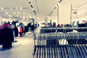 A clothing store interior featuring rows of metal clothing racks filled with hangers. The background is softly blurred, showcasing more clothing displays and bright ceiling lights, creating a spacious and modern retail environment.