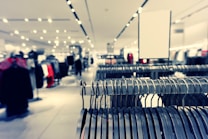 A clothing store interior featuring rows of metal clothing racks filled with hangers. The background is softly blurred, showcasing more clothing displays and bright ceiling lights, creating a spacious and modern retail environment.