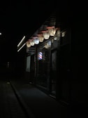 A dimly lit street scene featuring a barber shop with illuminated 'Barber' signage. The shop has a classic barber pole visible and other dimly lit features around it. The surrounding area looks dark and somewhat deserted.