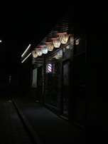 A dimly lit street scene featuring a barber shop with illuminated 'Barber' signage. The shop has a classic barber pole visible and other dimly lit features around it. The surrounding area looks dark and somewhat deserted.