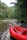 A red inflatable raft is positioned on a gently flowing river surrounded by dense forest. The lush green foliage of trees is abundant, with some branches extending over the water. Rocks line the edge of the river, enhancing the natural and untouched ambiance.
