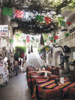 A charming restaurant interior with colorful tablecloths and rustic wooden chairs arranged in a dining area. Brightly colored papel picado flags hang overhead, adding a festive touch. The ceiling is adorned with greenery, giving a natural and inviting ambiance. A white staircase leads to an upper level, flanked by decorative white stucco walls and arches. Various clothing items hang on display to the left, and people are casually browsing.