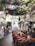 A charming restaurant interior with colorful tablecloths and rustic wooden chairs arranged in a dining area. Brightly colored papel picado flags hang overhead, adding a festive touch. The ceiling is adorned with greenery, giving a natural and inviting ambiance. A white staircase leads to an upper level, flanked by decorative white stucco walls and arches. Various clothing items hang on display to the left, and people are casually browsing.