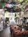 A charming restaurant interior with colorful tablecloths and rustic wooden chairs arranged in a dining area. Brightly colored papel picado flags hang overhead, adding a festive touch. The ceiling is adorned with greenery, giving a natural and inviting ambiance. A white staircase leads to an upper level, flanked by decorative white stucco walls and arches. Various clothing items hang on display to the left, and people are casually browsing.